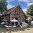 Flying Barista Ein rotes Fachwerkhaus mit Sitzgelegenheiten im Freien und Sonnenschirmen unter blauem Himmel.A red half-timbered house with outdoor seating and parasols under a blue sky.Et rødt bindingsværkshus med udendørs siddepladser og parasoller under en blå himmel.Een rood vakwerkhuis met buitenzitjes en parasols onder een blauwe hemel.