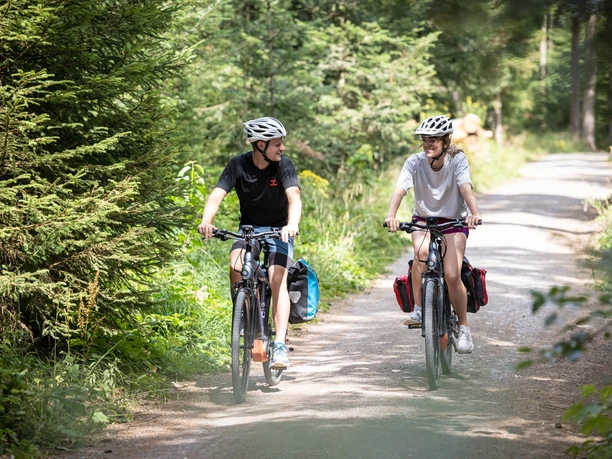 Ein junger Mann und eine junge Frau, beide fahren Rad auf einem Feldweg und lachen sich an.