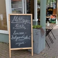 Tafel vor einem Geschäft wirbt mit "Mittagstisch: Linsen, Bulgursalat, vegan" neben Gemüsekisten.