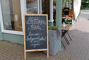 Unverpacktladen und Café PurProdukt Tafel vor einem Geschäft wirbt mit "Mittagstisch: Linsen, Bulgursalat, vegan" neben Gemüsekisten.