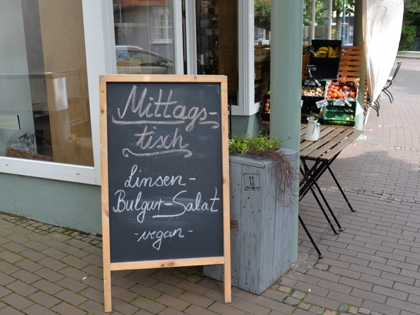 Unverpacktladen und Café PurProdukt Tafel vor einem Geschäft wirbt mit "Mittagstisch: Linsen, Bulgursalat, vegan" neben Gemüsekisten.