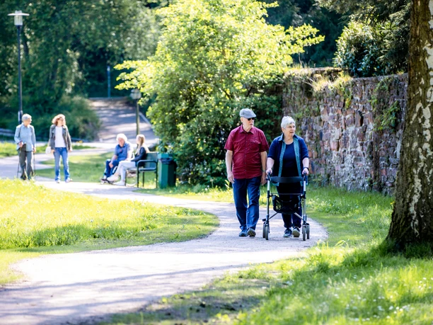 Bergischer Komfortspazierweg Ein älteres Paar geht mit einem Rollator in einem sonnigen Park, weitere Personen im Hintergrund.