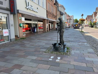 Eine Bronzestatue eines Glasbläsers steht auf einem steinernen Gehweg in einer Einkaufsstraße.