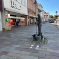 Eine Bronzestatue eines Glasbläsers steht auf einem steinernen Gehweg in einer Einkaufsstraße.