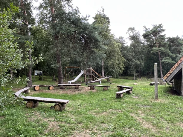 Spielplatz im Wald mit Klettergerüst, Rutsche und Sitzbänken aus Holz, umgeben von Bäumen.