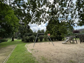 Spielplatz mit Schaukeln und Rutsche, umgeben von Bäumen auf einem Mehrgenerationenplatz.