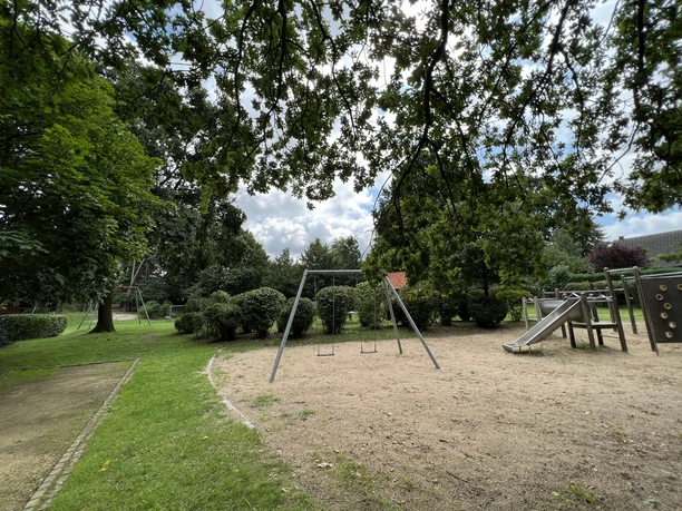 A playground with swings and a slide, surrounded by trees in a multigenerational park.