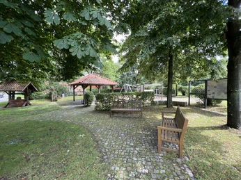 Ein Mehrgenerationenplatz mit breiten Gehwegen, Sitzbänken und einem überdachten Ruhebereich unter schattigen Bäumen.