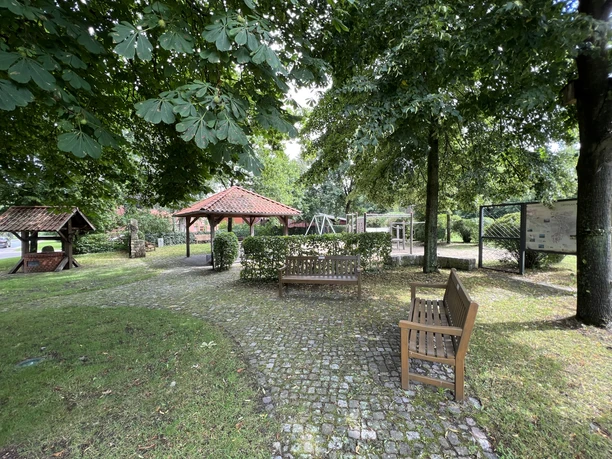 Ein Mehrgenerationenplatz mit breiten Gehwegen, Sitzbänken und einem überdachten Ruhebereich unter schattigen Bäumen.