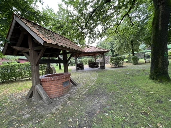 Ein gepflasterter Platz mit überdachtem Brunnen, umgeben von Bäumen und Sitzgelegenheiten im Freien.