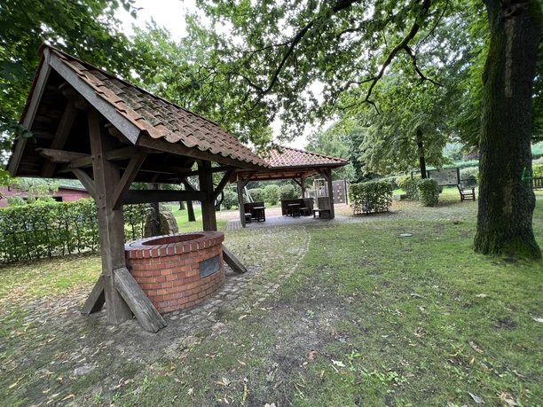 Ein gepflasterter Platz mit überdachtem Brunnen, umgeben von Bäumen und Sitzgelegenheiten im Freien.