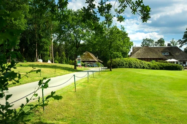 artland-golfclub Weg führt zu einem reetgedeckten Clubhaus im Artland Golfclub, umgeben von grüner Landschaft.