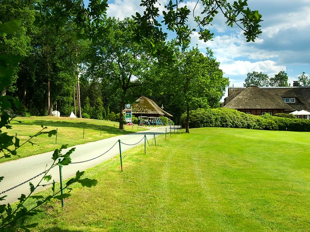 artland-golfclub Weg führt zu einem reetgedeckten Clubhaus im Artland Golfclub, umgeben von grüner Landschaft.