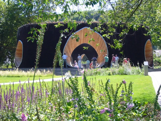 SoleArena Eine kreisförmige Holzkonstruktion umgeben von grüner Natur lädt Menschen zum Verweilen ein.A circular wooden construction surrounded by green nature invites people to linger.En cirkulær trækonstruktion omgivet af grøn natur inviterer folk til at blive hængende.Een ronde houten constructie omgeven door groene natuur nodigt mensen uit om te blijven hangen.