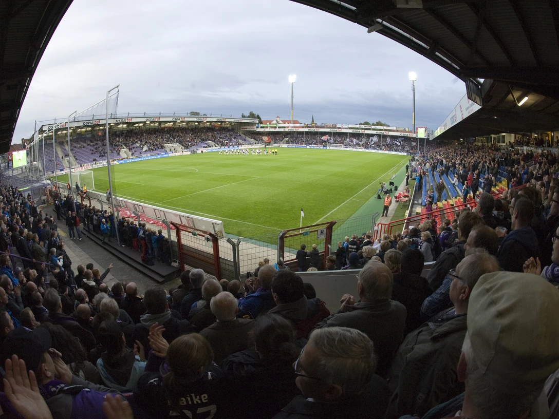 Fußballstadion bei Abendlicht mit vollen Tribünen und beleuchtetem Spielfeld, Fans jubeln lebhaft.