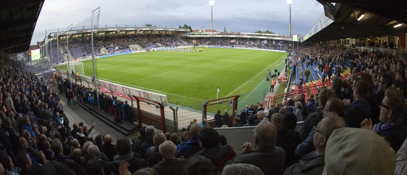 Fußballstadion bei Abendlicht mit vollen Tribünen und beleuchtetem Spielfeld, Fans jubeln lebhaft.