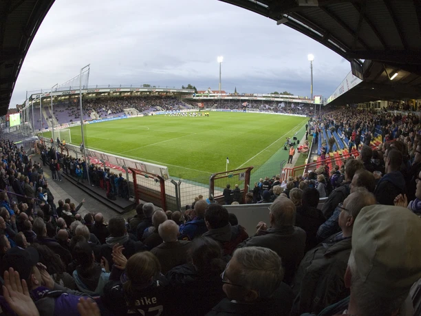 A soccer stadium in the evening light, with packed stands and a lit-up field; fans are cheering enthusiastically.