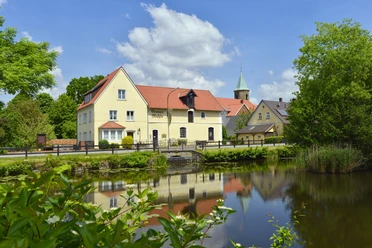 Venner Mühleninsel Historisches Gasthaus mit rotem Dach spiegelt sich in ruhigem Teich, umgeben von grüner Natur.