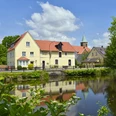 Venner Mühleninsel Historisches Gasthaus mit rotem Dach spiegelt sich in ruhigem Teich, umgeben von grüner Natur.