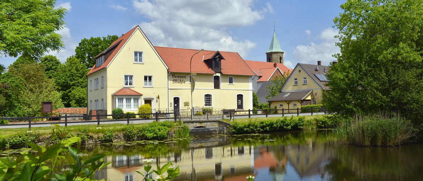 Venner Mühleninsel Historisches Gasthaus mit rotem Dach spiegelt sich in ruhigem Teich, umgeben von grüner Natur.