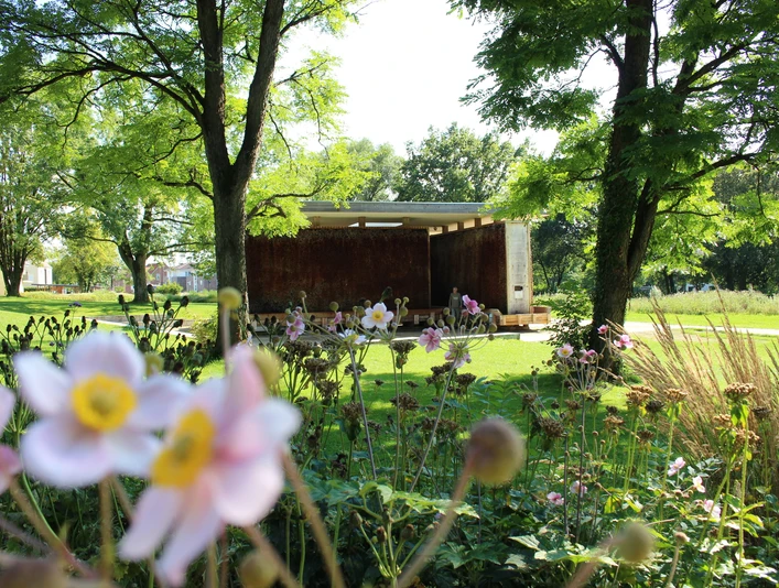Der SolePavillon im Kurpark in Bad Laer Rostfarbene Stahlwand in einem sonnigen Park, umgeben von blühenden Blumen und Bäumen.Rust-colored steel wall in a sunny park, surrounded by blossoming flowers and trees.Rustfarvet stålvæg i en solrig park, omgivet af blomstrende blomster og træer.Roestkleurige stalen muur in een zonnig park, omringd door bloeiende bloemen en bomen.