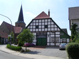 Gildehaus mit Kirche in Bissendorf Fachwerkhaus mit grünen Akzenten, dahinter Kirchturm, am Straßenrand mit Blumen und geparktem Auto.