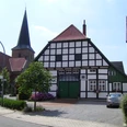 Gildehaus mit Kirche in Bissendorf Fachwerkhaus mit grünen Akzenten, dahinter Kirchturm, am Straßenrand mit Blumen und geparktem Auto.