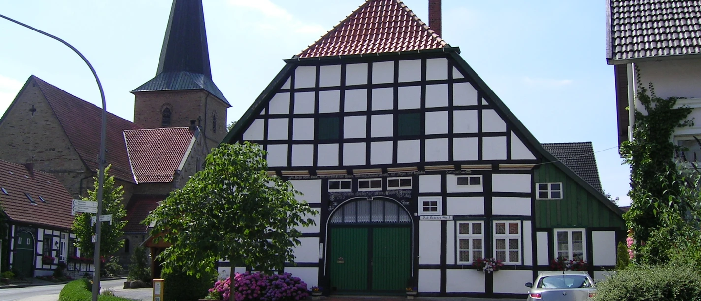 Gildehaus mit Kirche in Bissendorf Fachwerkhaus mit grünen Akzenten, dahinter Kirchturm, am Straßenrand mit Blumen und geparktem Auto.