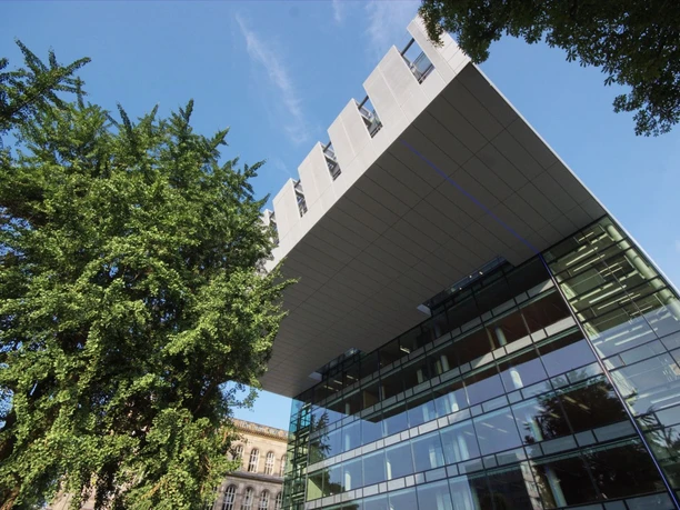 Blick von unten hoch zu einem modernen Gebäude in C-Form mit Glasfront, umgeben von grünen Bäumen.
