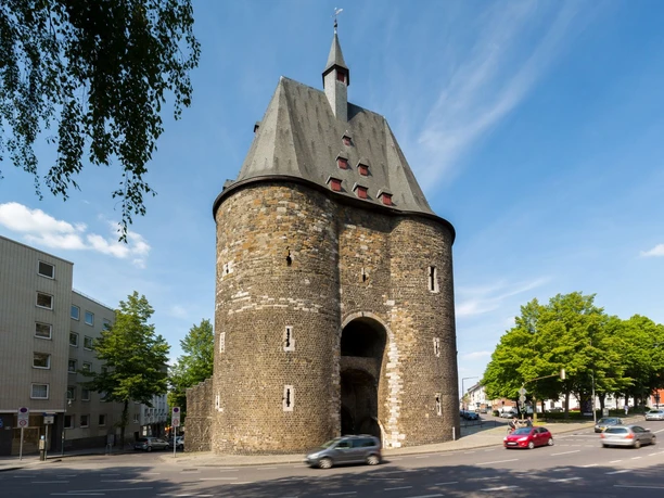Marschiertor Aachen Mittelalterliches Stadttor aus Backstein, mit runden Türmen und kleinen Gauben. Im Vordergrund befinden sich Bäume und eine Straße.
