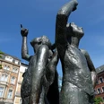 Drei Bronze-Figuren stehen mit dem Rücken aneinander und heben den kleinen Finger in die Luft.