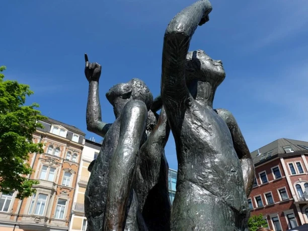 Drei Bronze-Figuren stehen mit dem Rücken aneinander und heben den kleinen Finger in die Luft.