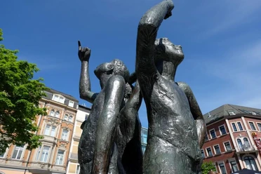 Klenkes Drei Bronze-Figuren stehen mit dem Rücken aneinander und heben den kleinen Finger in die Luft.