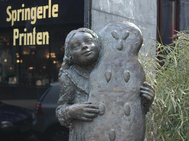Bronze-Figur eines Mädchens, das eine fast körpergroße, mit Mandeln verzierte Printe, ebenfalls aus Bronze, in den Händen hält.