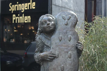 Printenmädchen Bronze-Figur eines Mädchens, das eine fast körpergroße, mit Mandeln verzierte Printe, ebenfalls aus Bronze, in den Händen hält.