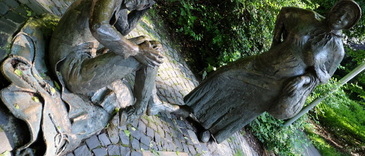 Zwei Bronze-Figuren nebeneinander. Die eine stellt eine Marktfrau mit Korb dar, die andere den Teufel mit Hörnern, sitzend und sich am Kopf kratzend. Im Boden davor ist eine Informationstafel eingelassen.