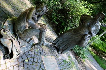 Teufel und Marktfrau Zwei Bronze-Figuren nebeneinander. Die eine stellt eine Marktfrau mit Korb dar, die andere den Teufel mit Hörnern, sitzend und sich am Kopf kratzend. Im Boden davor ist eine Informationstafel eingelassen.