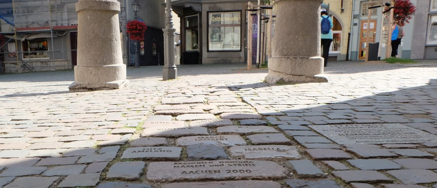 Weg aus Pflastersteinen, in die verschiedene Daten eingemeißelt sind. Zwei Poller begrenzen den Weg.