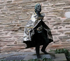 Die Bronzestatue zeigt einen gekrönten Mann im langen Umhang, vor einer Steinwand gehend.