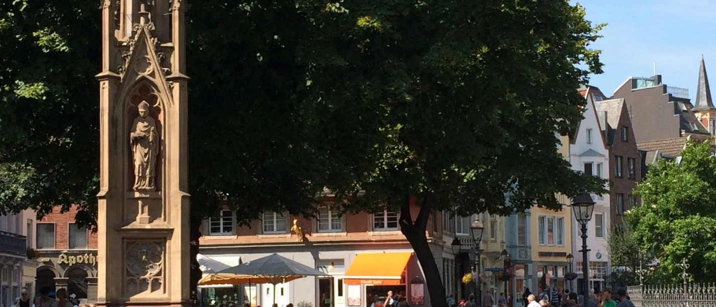 Gotischer Brunnen unter einem großen Baum. Um den Brunnen herum stehen Sitzmöglichkeiten und Menschen flanieren oder sitzen in einer Außengastronomie.
