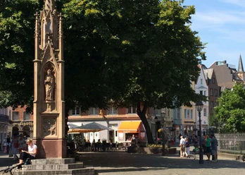 Marienbrunnen Gotischer Brunnen unter einem großen Baum. Um den Brunnen herum stehen Sitzmöglichkeiten und Menschen flanieren oder sitzen in einer Außengastronomie.