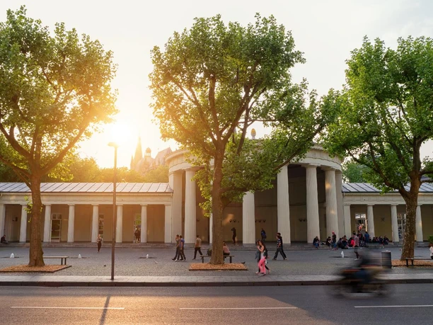 Elisenbrunnen Gebäudekomplex des Elisenbrunnens mit Bäumen im Vordergrund und dem Sonnenuntergang dahinter.