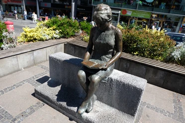 Eine Bronze-Statue sitze lesend auf einer Bank umgeben von Blumenkübeln.