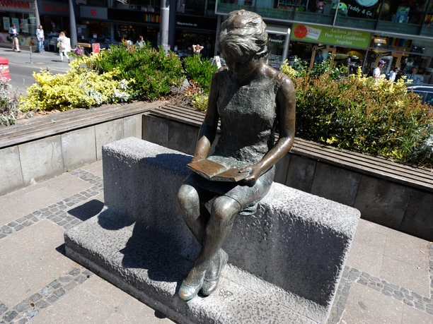 Eine Bronze-Statue sitze lesend auf einer Bank umgeben von Blumenkübeln.