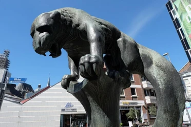 Bahkauv Bronzeskulptur eines löwenartigen Wesens auf einem Sockel in urbaner Umgebung.
