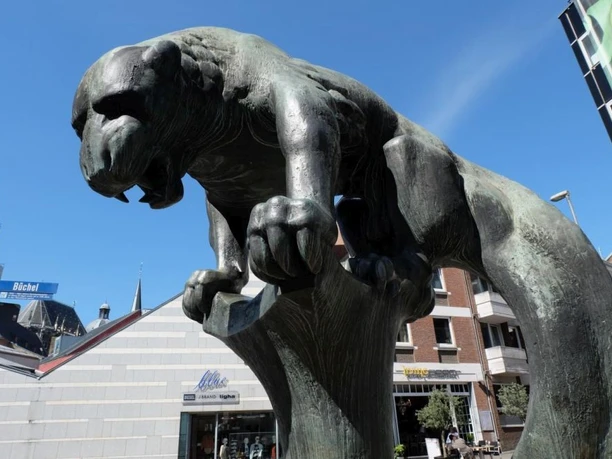Bahkauv Bronzeskulptur eines löwenartigen Wesens auf einem Sockel in urbaner Umgebung.