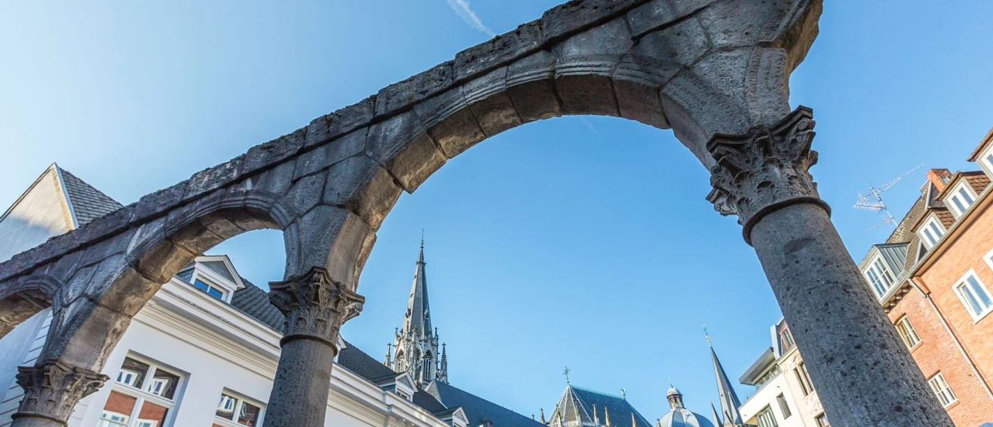 Steinerne Torbögen aus der Römerzeit stehen unter blauem Himmel im Hof. Im Hintergrund ragt der Aachener Dom empor.