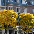 Hühnerdieb Vor einem herbstlichen Baum mit gelben Blättern steht eine Statue von einem kleinen Jungen mit einem Huhn auf dem Arm auf einem sonnigen Platz