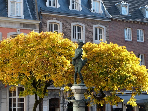 Hühnerdieb Vor einem herbstlichen Baum mit gelben Blättern steht eine Statue von einem kleinen Jungen mit einem Huhn auf dem Arm auf einem sonnigen Platz