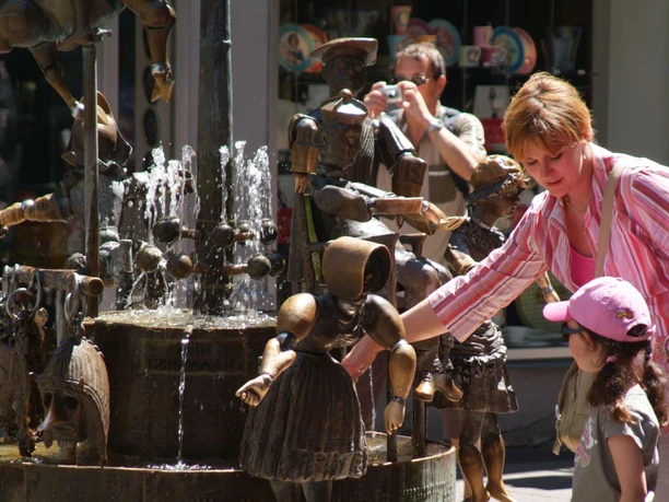 Brunnen mit verschiedenen Figuren aus Bronze, darunter Personen in altertümlicher Kleidung. Eine Touristin betrachtet den Brunnen gerade.